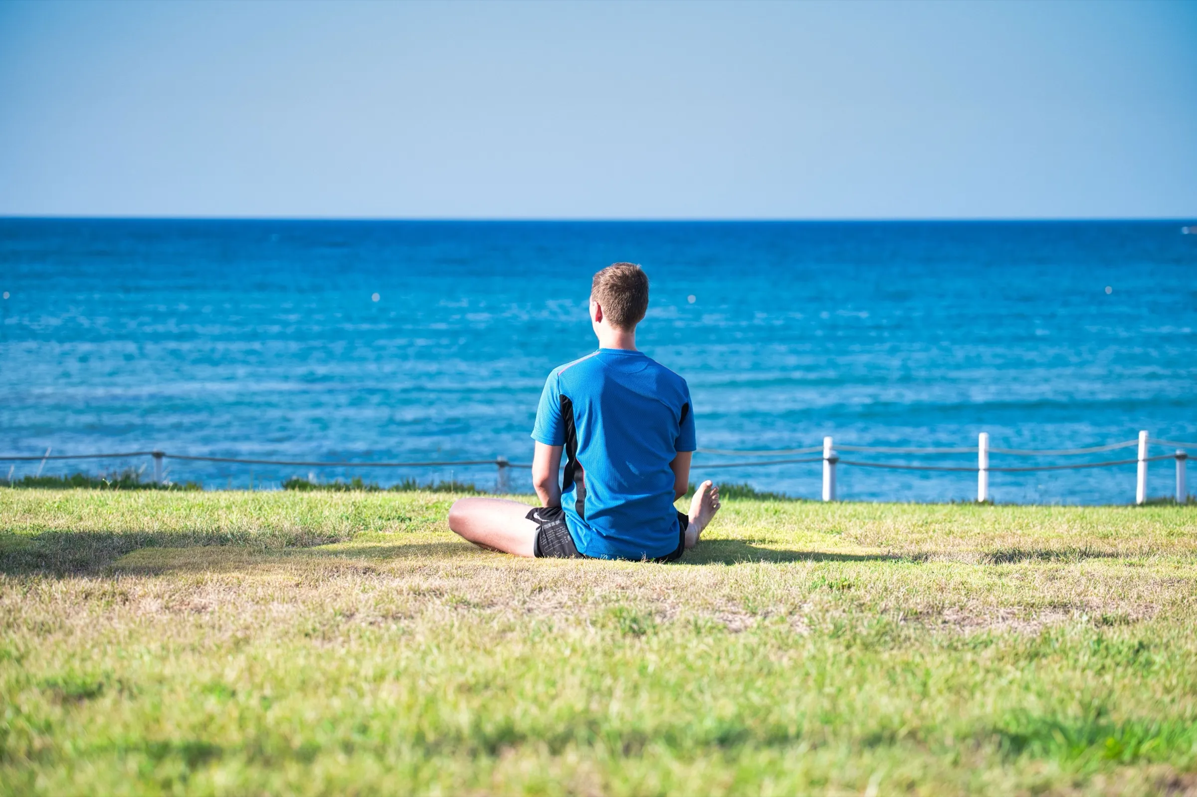 Eine Person sitzt aufrecht in Meditationshaltung im Gras am Meer und blickt ruhig aufs Wasser